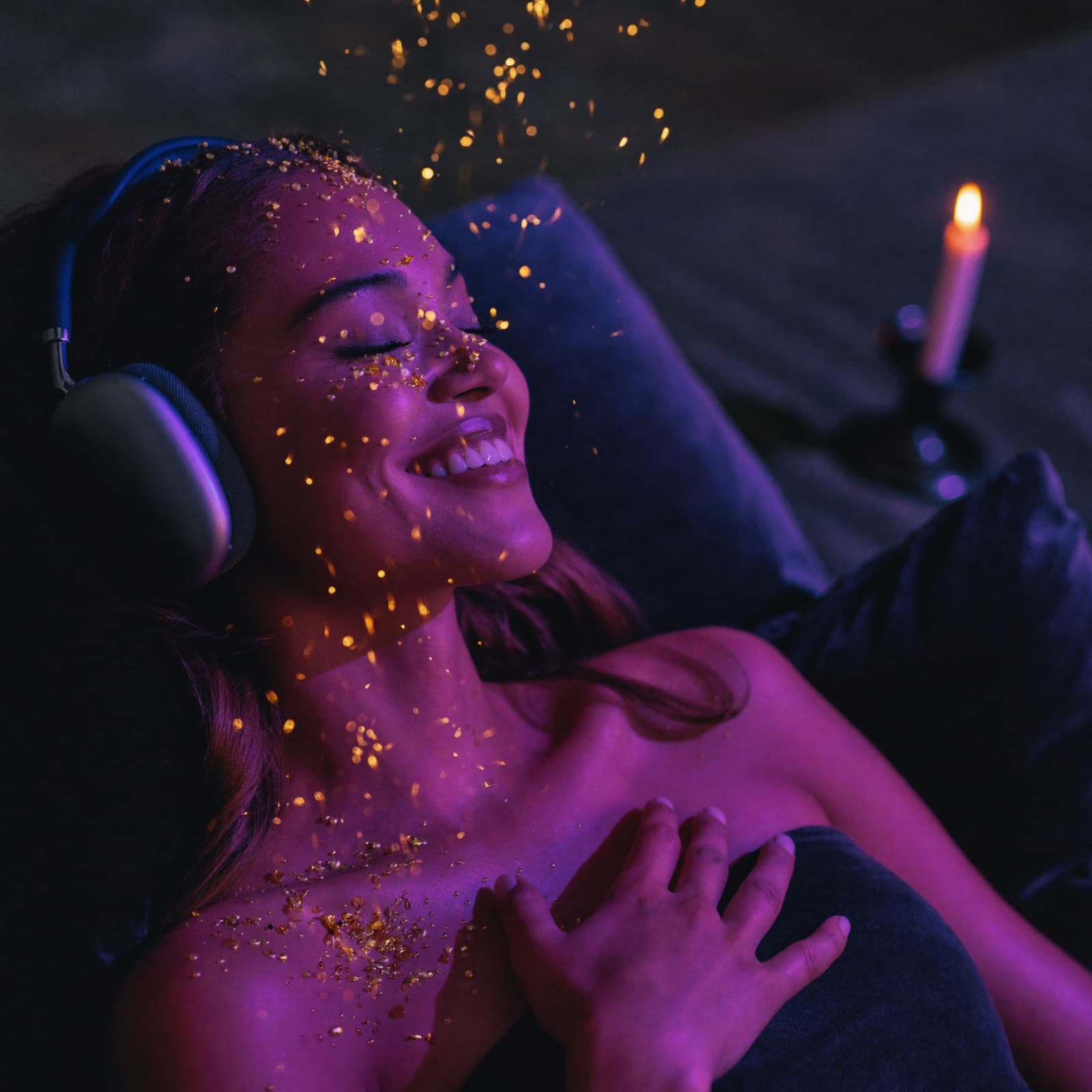 A woman reclined with headphones on, eyes closed and smiling, golden sparks drifting over her in deep violet candlelight